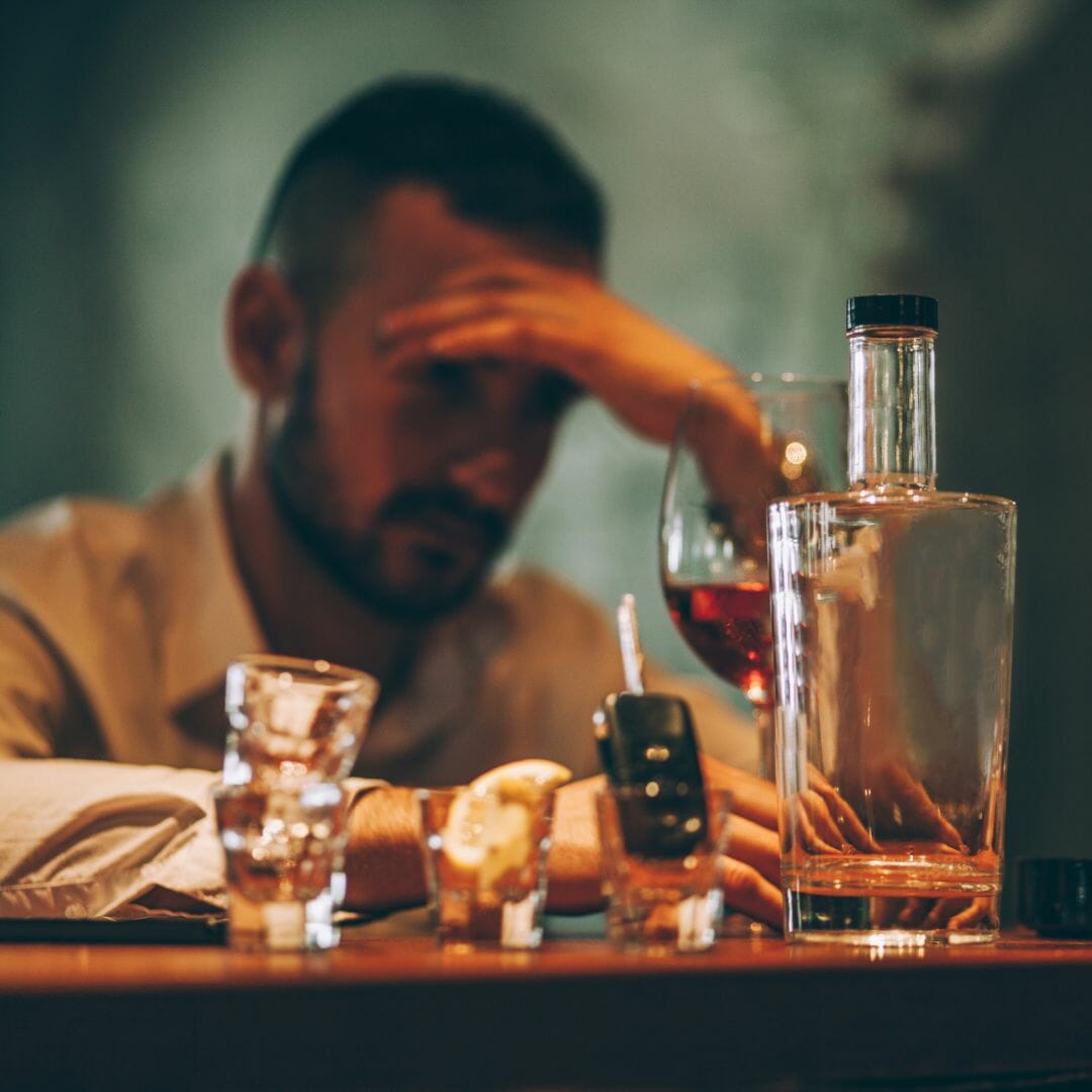 man sitting depressed with a glass of wine and an empty liquor bottle, shot glasses and his car key in a shot glass